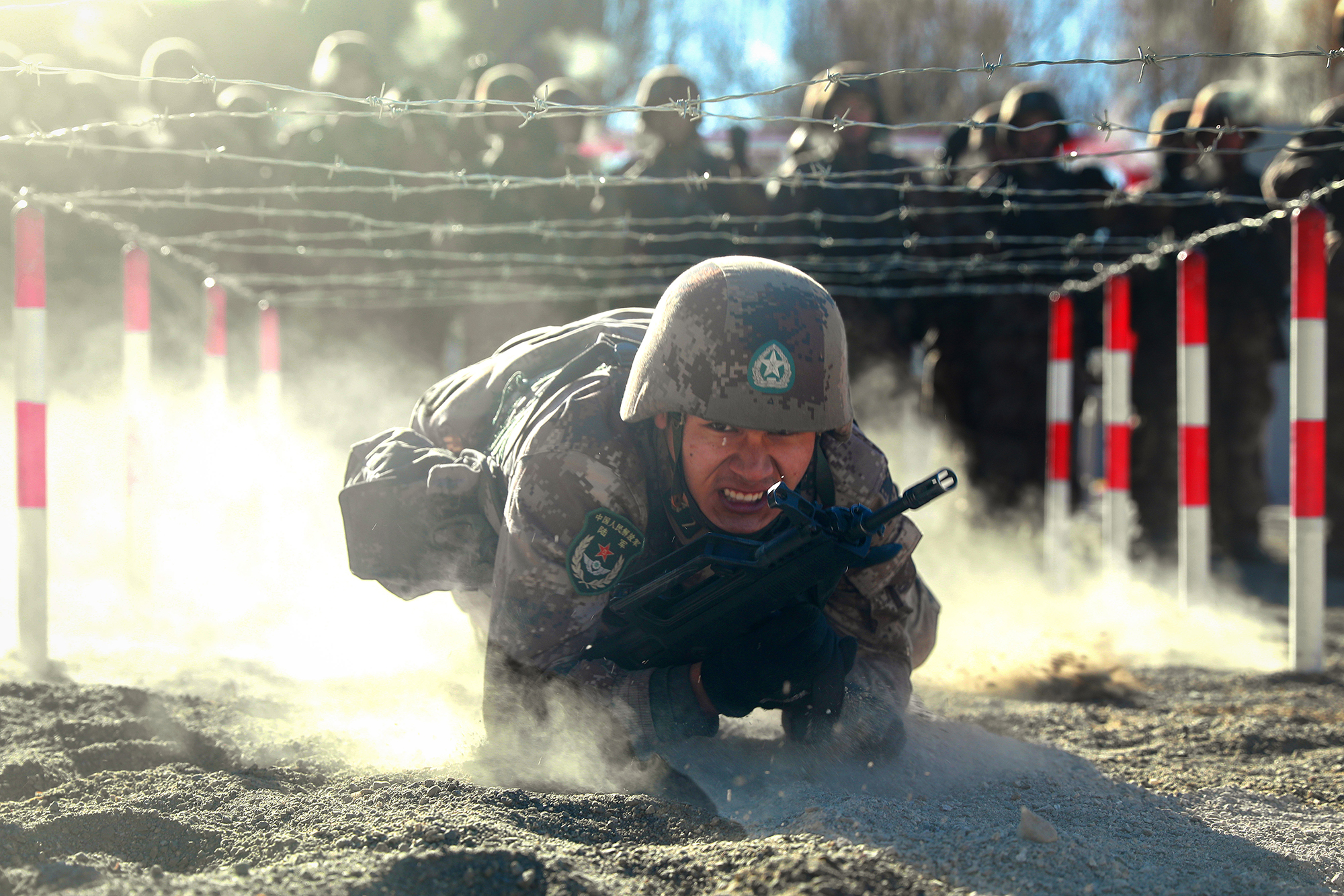 A Chinese soldier low-crawls in the dirt under barbed wire during training.